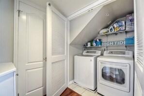 A laundry room with two front-loading washing machines, a dryer, and shelves with laundry supplies.