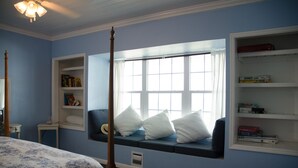 A bedroom with a large bed, a window seat with pillows, a bookshelf, and a small table.