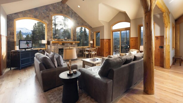 A spacious living area with a stone fireplace, large windows offering mountain views, a cozy seating area, and a wooden ceiling beam. - Moosehead Lodge: Clear Mountain Views, Hot Tub & Game Room (Breckenridge)