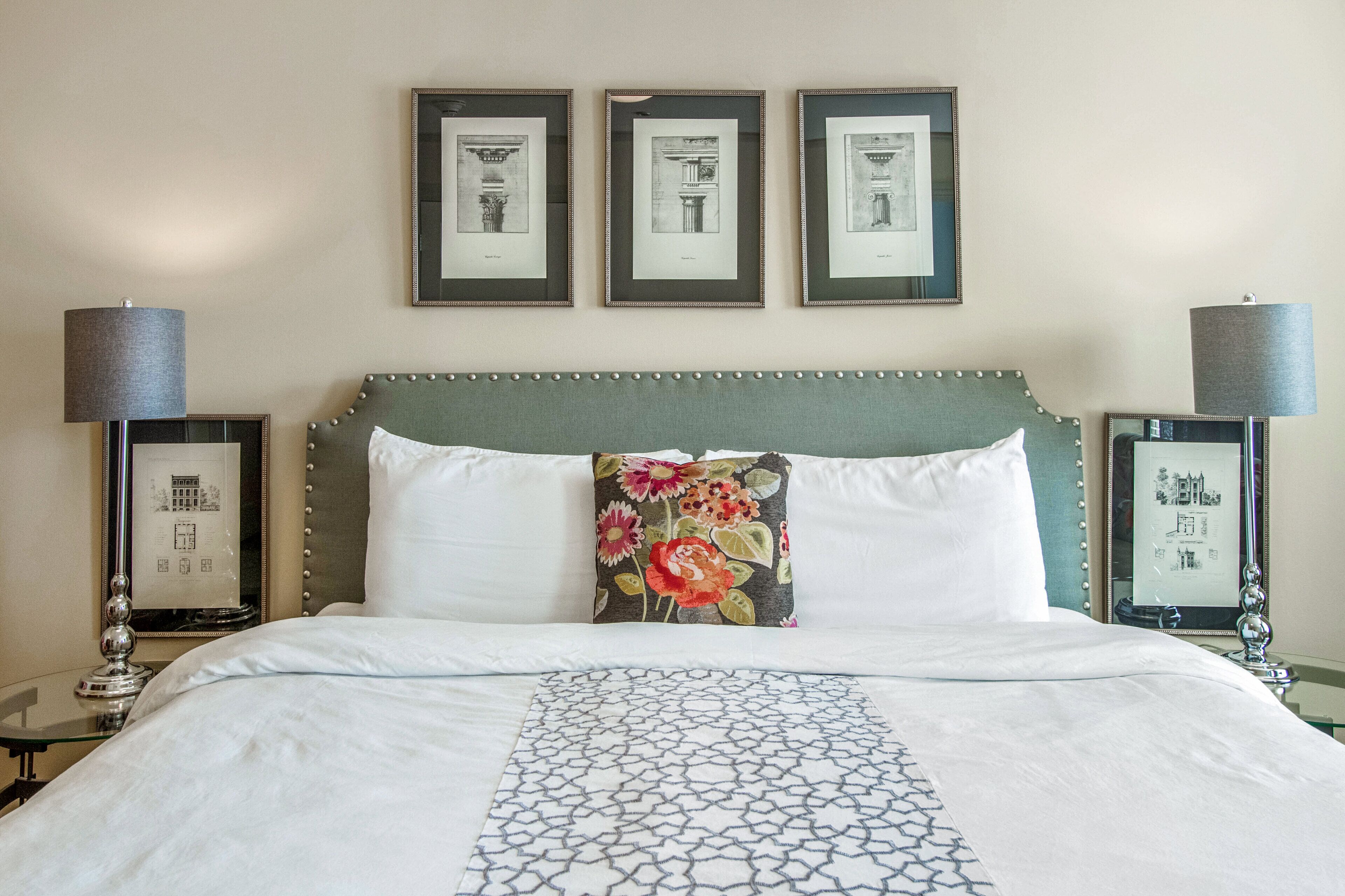 A neatly made bed with a patterned bedspread, a decorative pillow, and a headboard with a beaded trim. There are three framed pictures on the wall above the bed.
