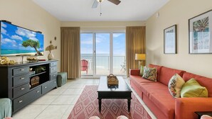 A living room with a television, a ceiling fan, a sofa, a coffee table, and a view of the beach through the sliding glass door. - Ariel Dunes - Amazing Gulf Views, Pools, Gym, Walk to Beach, On-Site Dining.  😎 (Miramar Beach)