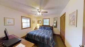Ein Schlafzimmer mit einem Bett, einem Schreibtisch mit Computer, einem Deckenventilator und zwei Fenstern.