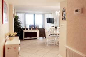 A modern living room with a dining area, a flat-screen TV mounted on the wall, and a large window with blinds.