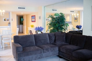 A living room with a dark grey sectional sofa, a dining area with white chairs, and a large green plant.