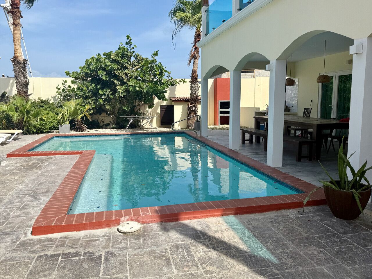 A rectangular swimming pool with a red brick border, surrounded by a patio area with stone tiles.