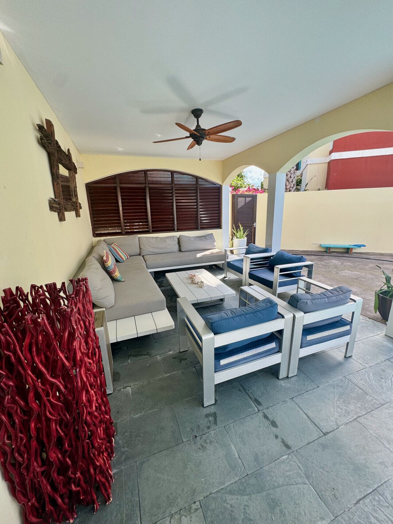Un espace patio aménagé avec un canapé, deux fauteuils et une table basse. On y trouve également un ventilateur de plafond, un banc en bois et un objet décoratif rouge.