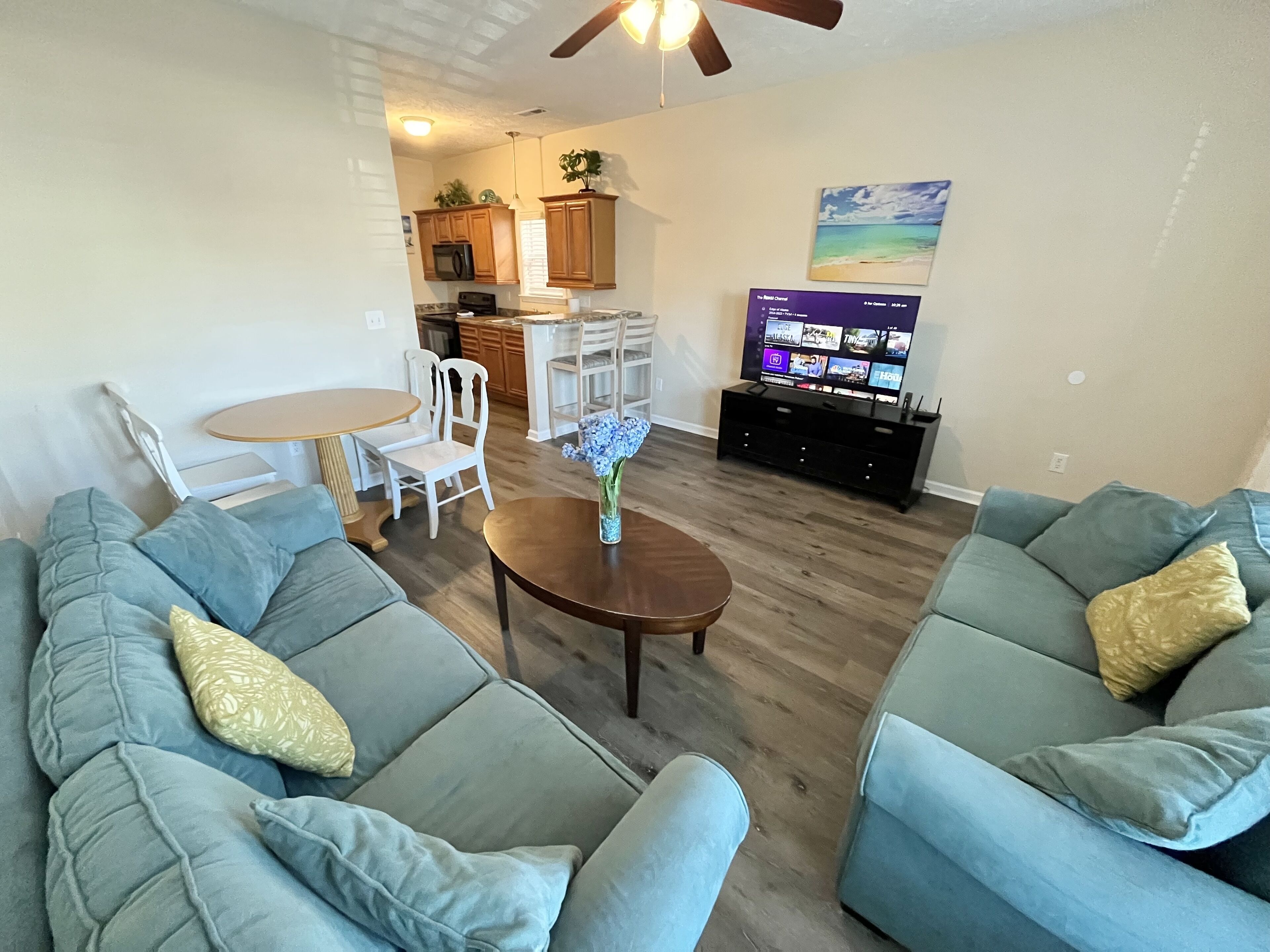 Ein Wohnzimmer mit zwei Sofas, einem Couchtisch und einem Fernseher. Im Hintergrund ist eine Küchenzeile mit Holzausbau und einer Mikrowelle zu sehen.