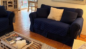 A cozy living room with a dark blue sofa, a wooden coffee table with magazines and candles, and a large window with a view of boats. - Highly Rated Cottage in Orleans, Cape Cod (Orleans)