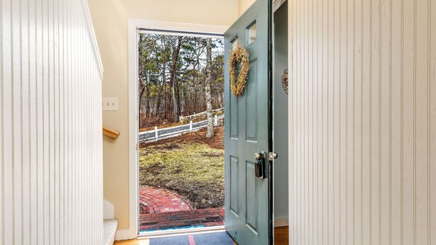 A wooden door with a wreath, a white door frame, and a view of a brick pathway leading to a white fence and trees. - Highly Rated Cottage in Orleans, Cape Cod (Orleans)