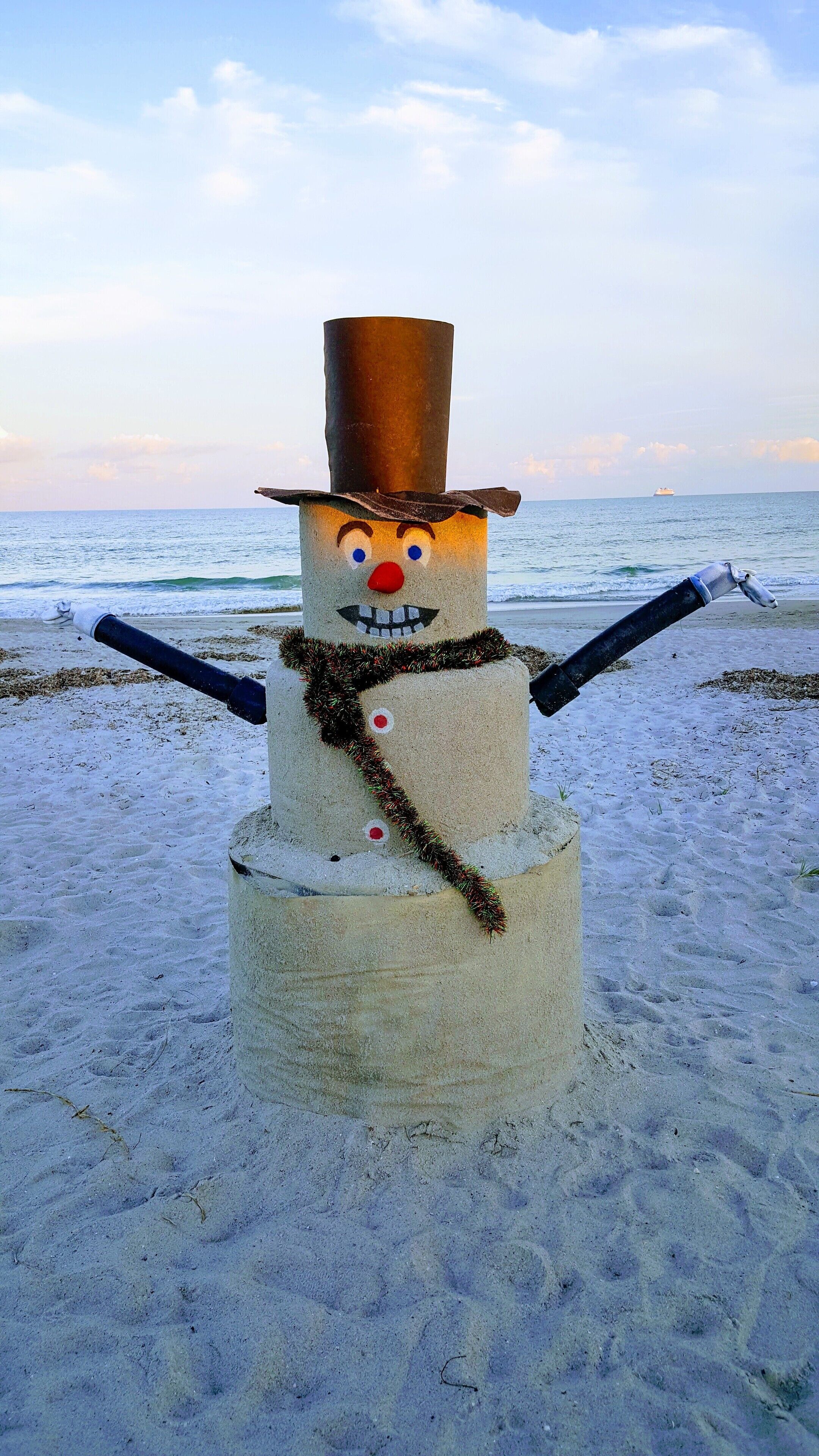Una escultura de muñeco de nieve en la playa, con un barco al fondo.