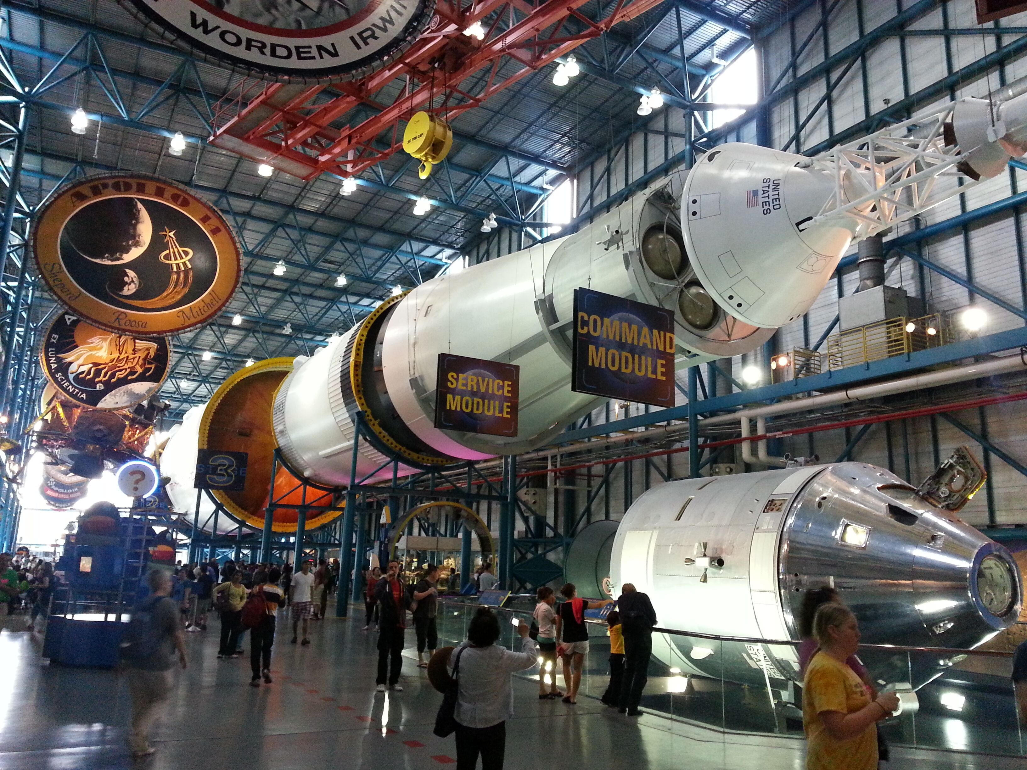 A large indoor exhibition hall with various spacecraft models on display, including a prominent Saturn V rocket.
