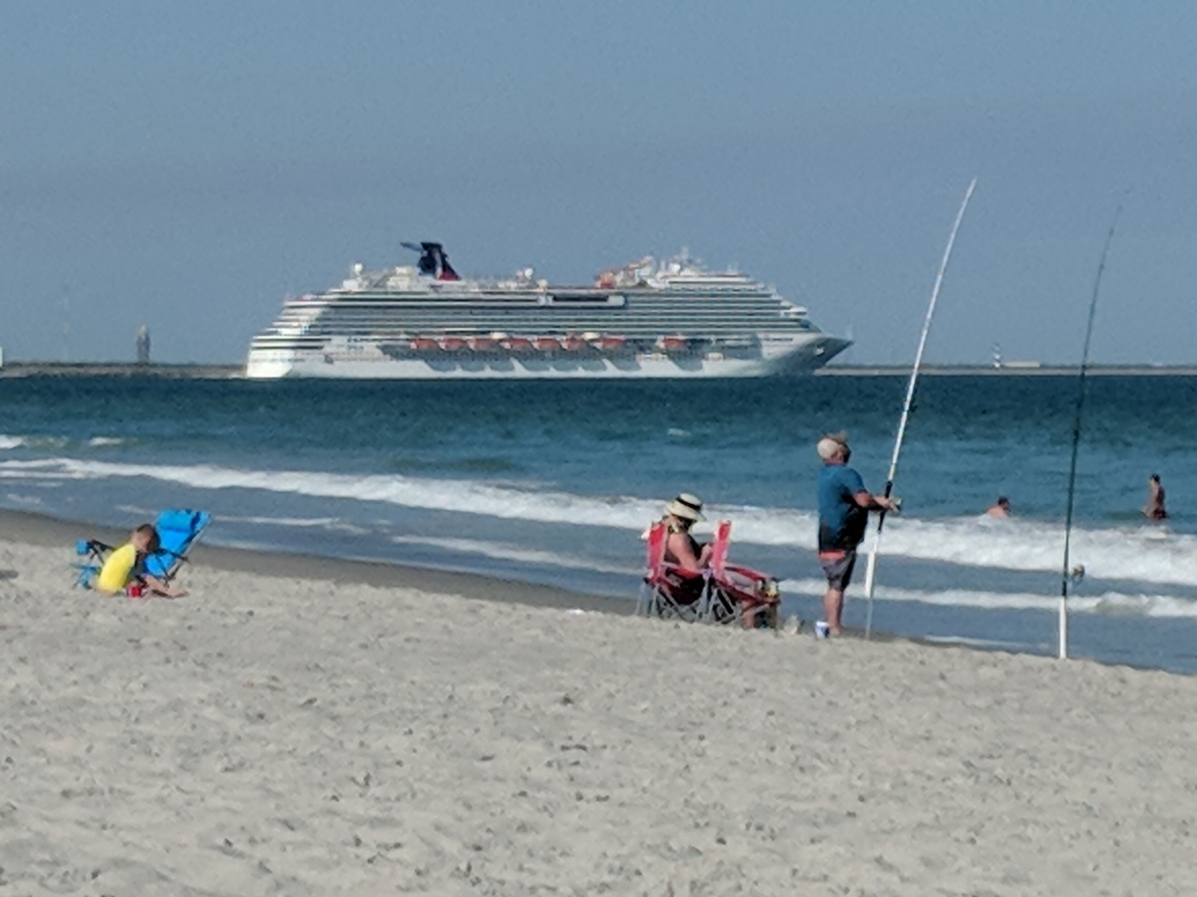 Una playa con un gran crucero al fondo, gente pescando y sillas de playa.