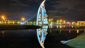 Ein modernes Gebäude mit markantem, gebogenem Design, das sich in ruhigem Wasser spiegelt, umgeben von einem gut beleuchteten Stadtgebiet.