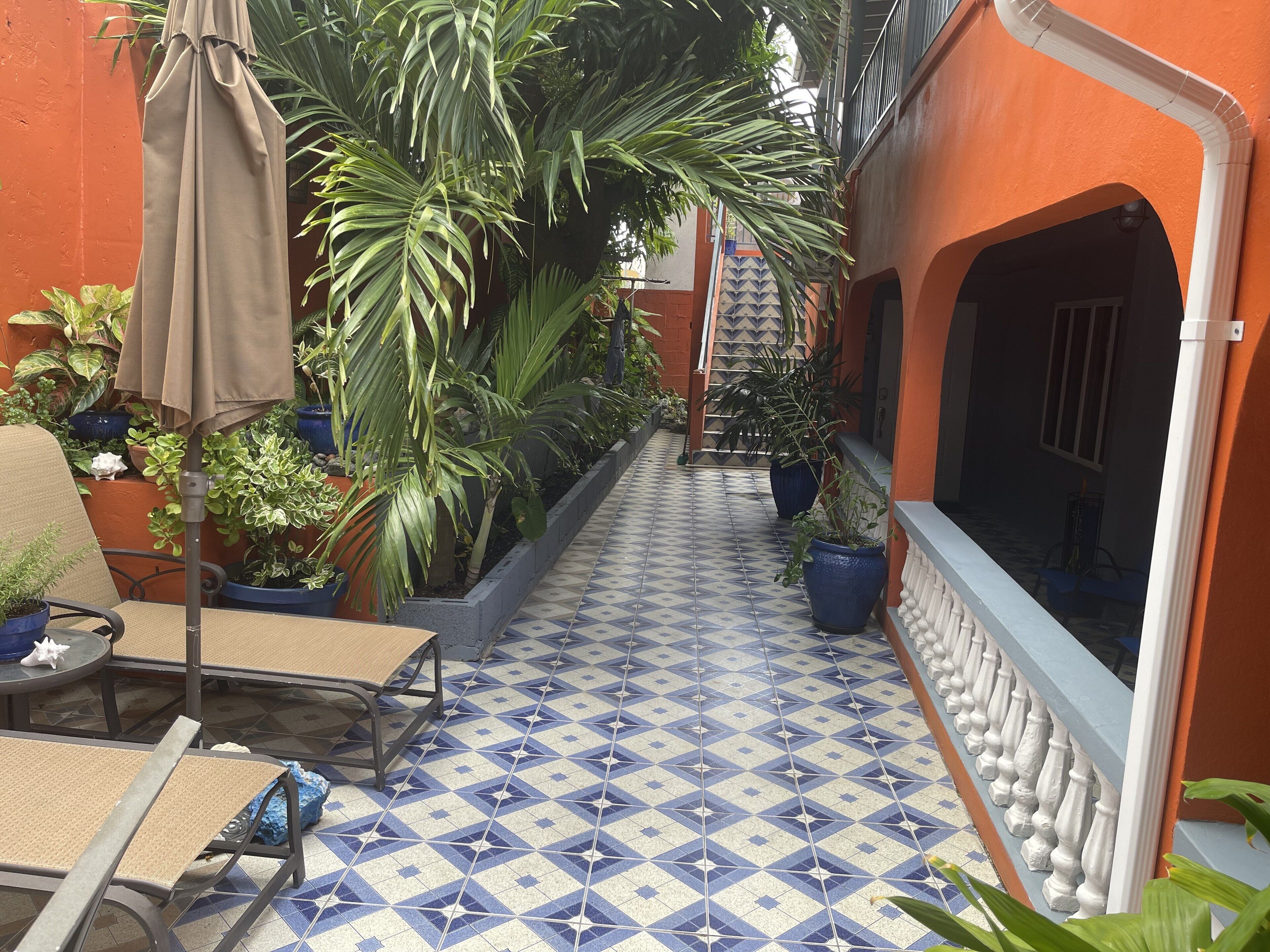 A narrow outdoor corridor with tiled flooring, potted plants, and outdoor seating.