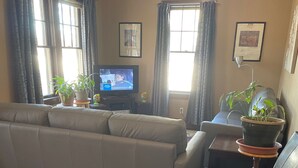 Ein Wohnzimmer mit einer Couch, einem Sessel, einem Fernseher und Blumentöpfen.