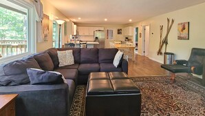 A modern living room with a dark brown sectional sofa, a black ottoman, and a patterned rug. A dining area with a wooden table and chairs is visible in the background. - Spacious 6BR - 5BA Ski Home on Pico Mountain - Minutes to Killington Mountain (Killington)