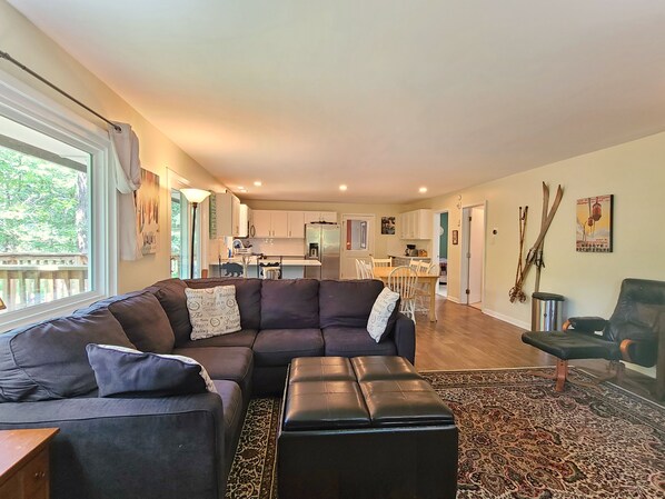 A modern living room with a dark brown sectional sofa, a black ottoman, and a patterned rug. A dining area with a wooden table and chairs is visible in the background.