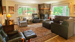 A cozy living room with a fireplace, leather sofas, a patterned rug, and large windows offering a view of the outdoors. - Spacious 6BR - 5BA Ski Home on Pico Mountain - Minutes to Killington Mountain (Killington)