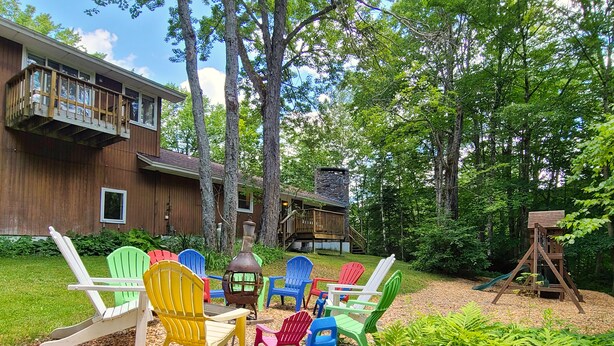 A backyard with a wooden deck, colorful Adirondack chairs, a stone fireplace, and a wooden swing set. - Spacious 6BR - 5BA Ski Home on Pico Mountain - Minutes to Killington Mountain (Killington)