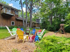 A backyard with a wooden deck, colorful Adirondack chairs, a stone fireplace, and a wooden swing set.