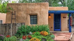 Un bâtiment rustique avec une porte bleue et une fenêtre, entouré d’un jardin où poussent différentes plantes et fleurs.