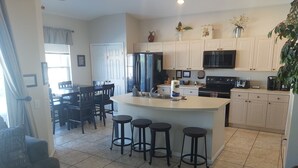 A modern kitchen with a central island, bar stools, a dining area with chairs, and a refrigerator. - Luxury Pool Home Steps to Disney, 2 Master Suites, Gated, Game Rm, BBQ Free WiFi (Kissimmee)