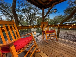 Una terraza de madera con dos mecedoras y una mesita, rodeada de árboles y luces colgantes.