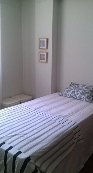 Uma cama pequena e bem arrumada com um edredom listrado e um travesseiro estampado com motivos florais. Duas quadros emolduradas estão penduradas na parede acima da cama.