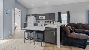 A modern kitchen with grey cabinets, a white island, and a microwave. There is a dark brown sofa and a window with blue curtains. - Recently renovated 5BR/4BA villa with private pool&spa located in Hampton Lakes. (Davenport)