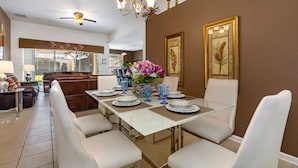 Un comedor con una mesa blanca dispuesta para seis personas, rodeada de sillas blancas. Hay un ventilador de techo arriba y una araña en la pared.