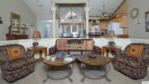 A spacious living room with a large sofa, two armchairs, and a coffee table. There is a dining area in the background with a chandelier.