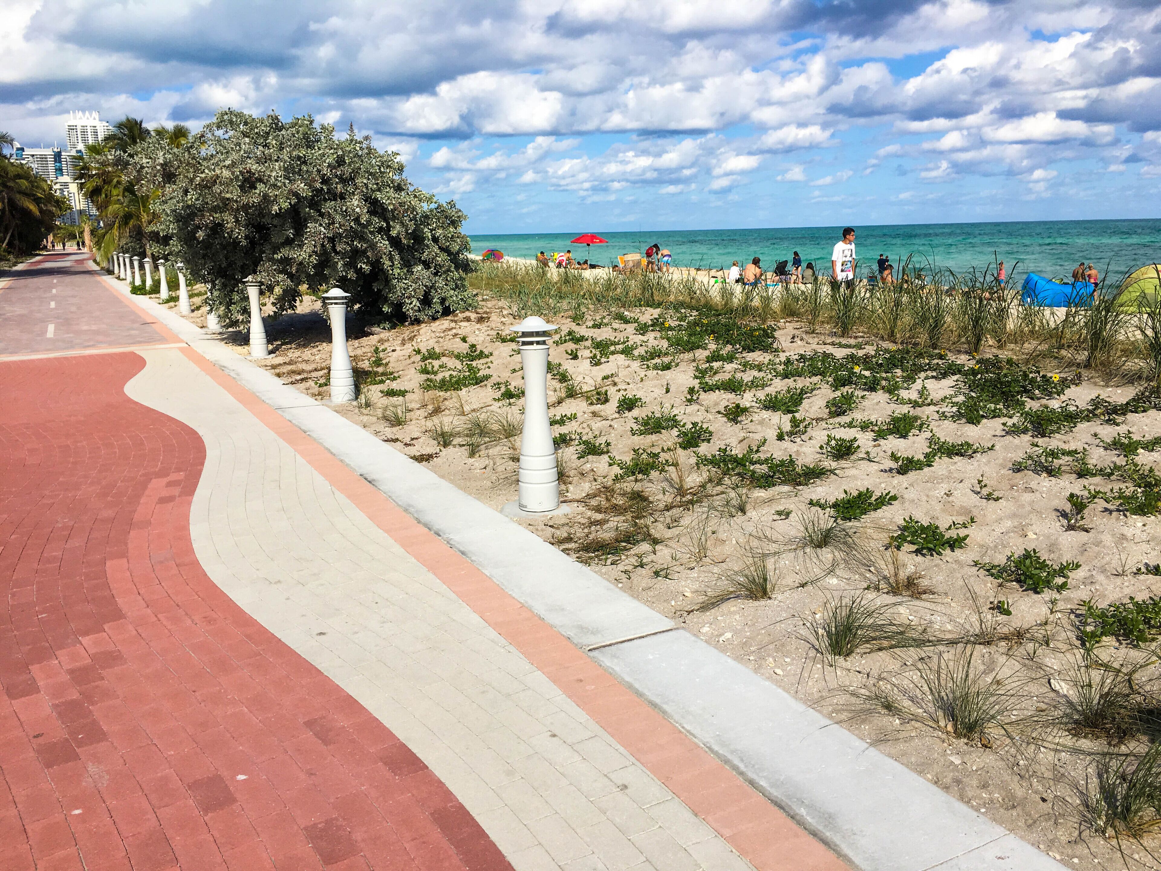 A paved walkway with bollards, a sandy beach area with sparse vegetation, and the ocean in the background.