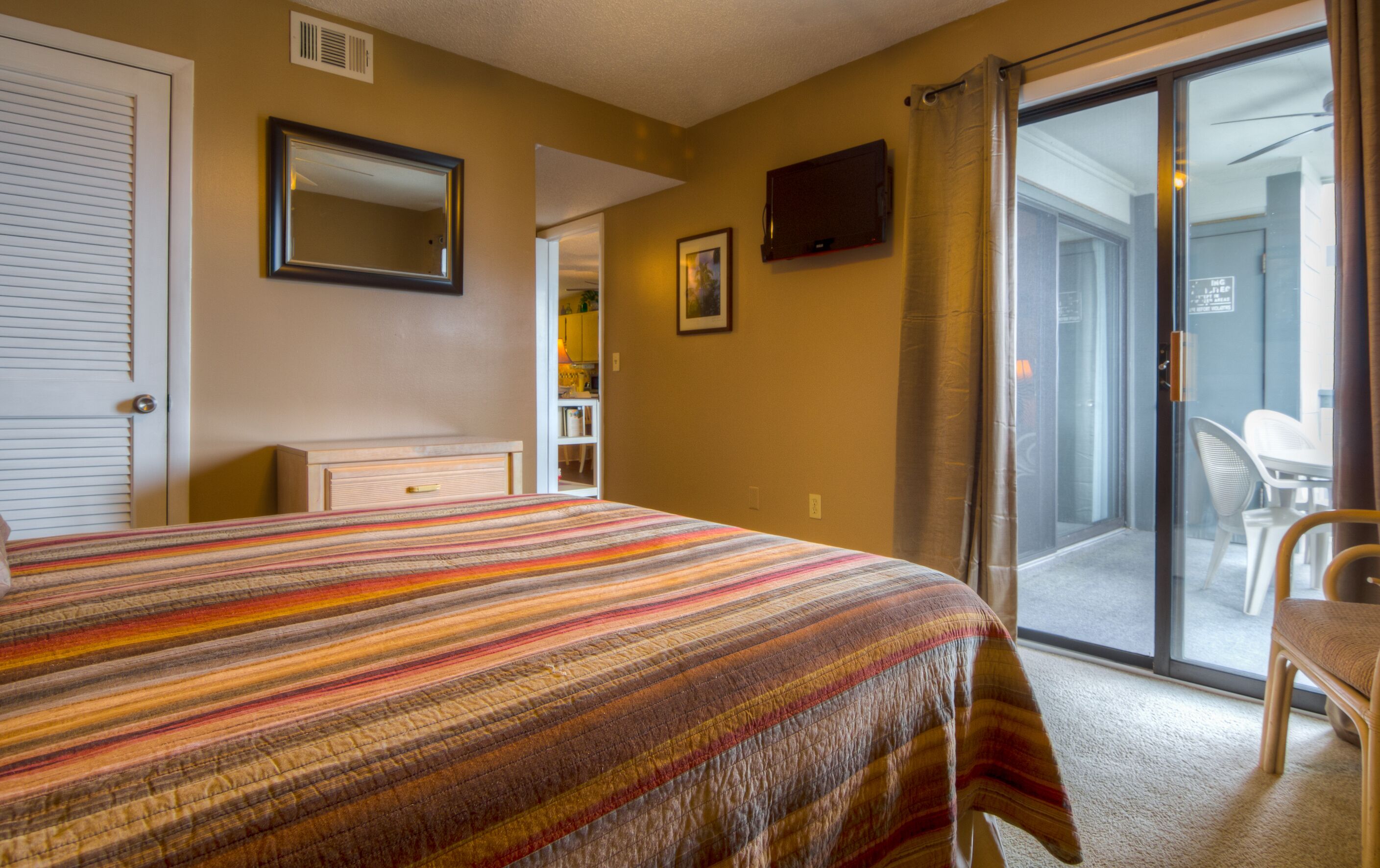 A hotel room with a bed, a sliding glass door to the balcony, a television on the wall, and a small table with a chair.