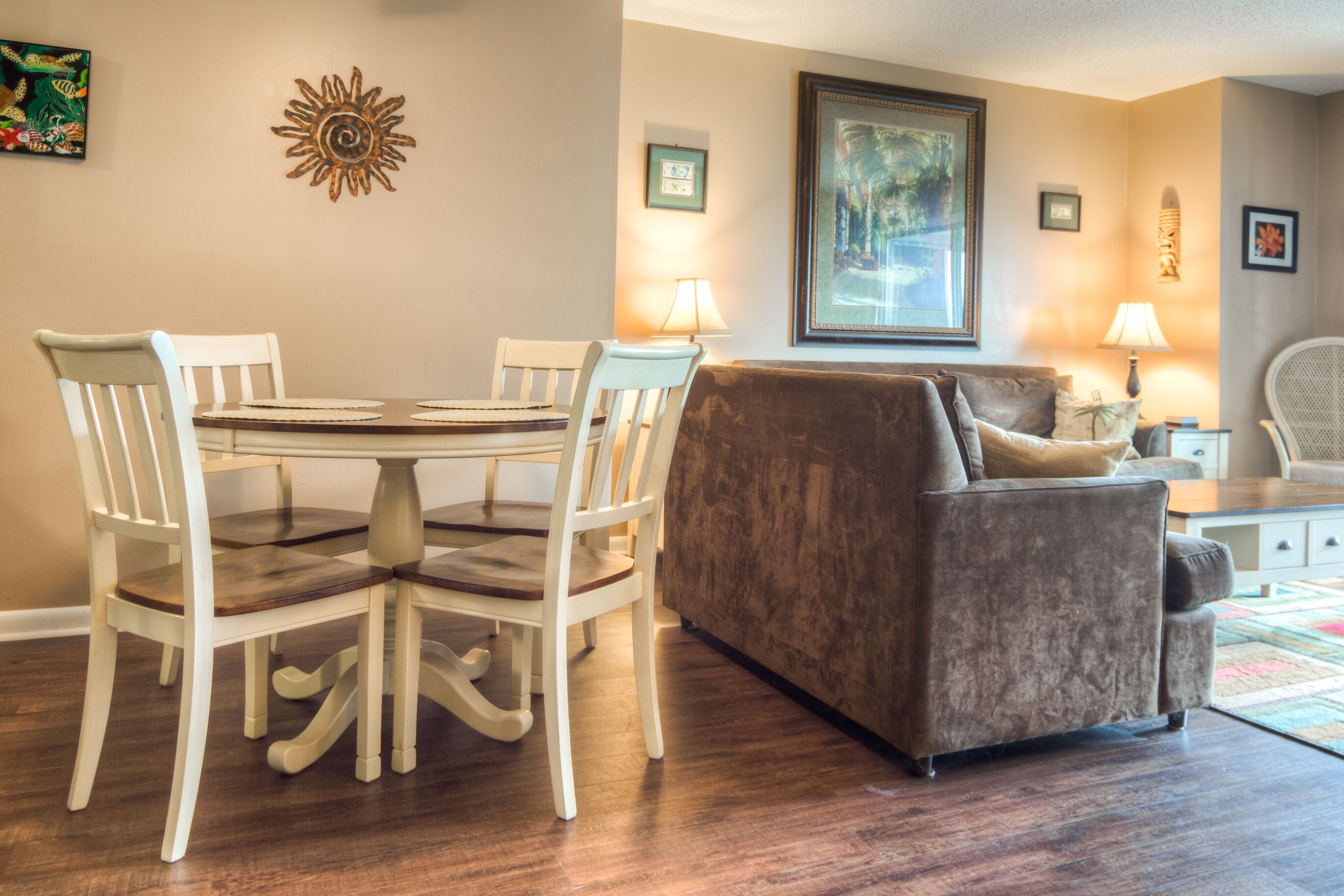 Een eetgedeelte met een ronde tafel en vier stoelen, een bruine bank en een lamp op een bijzettafeltje.