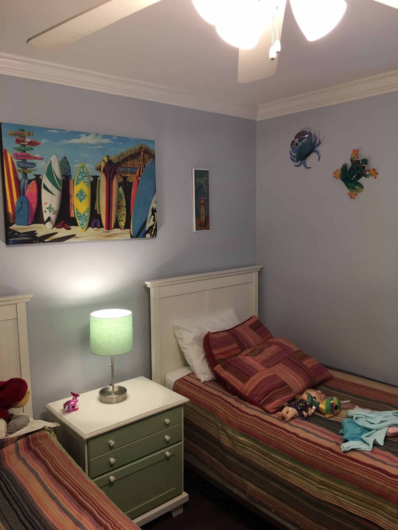 A bedroom with a bed, a nightstand, a lamp, and a wall with surfboards and a frog decoration.