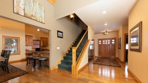 A spacious interior with a wooden staircase, a kitchen area, and a dining space with a view of trees.