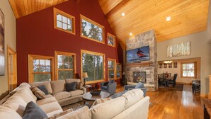 A spacious living room with a large sectional sofa, a stone fireplace, and multiple windows offering views of the outdoors.