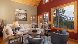 A cozy living room with a large window offering a mountain view, a beige sectional sofa, a round coffee table, and a grey armchair.
