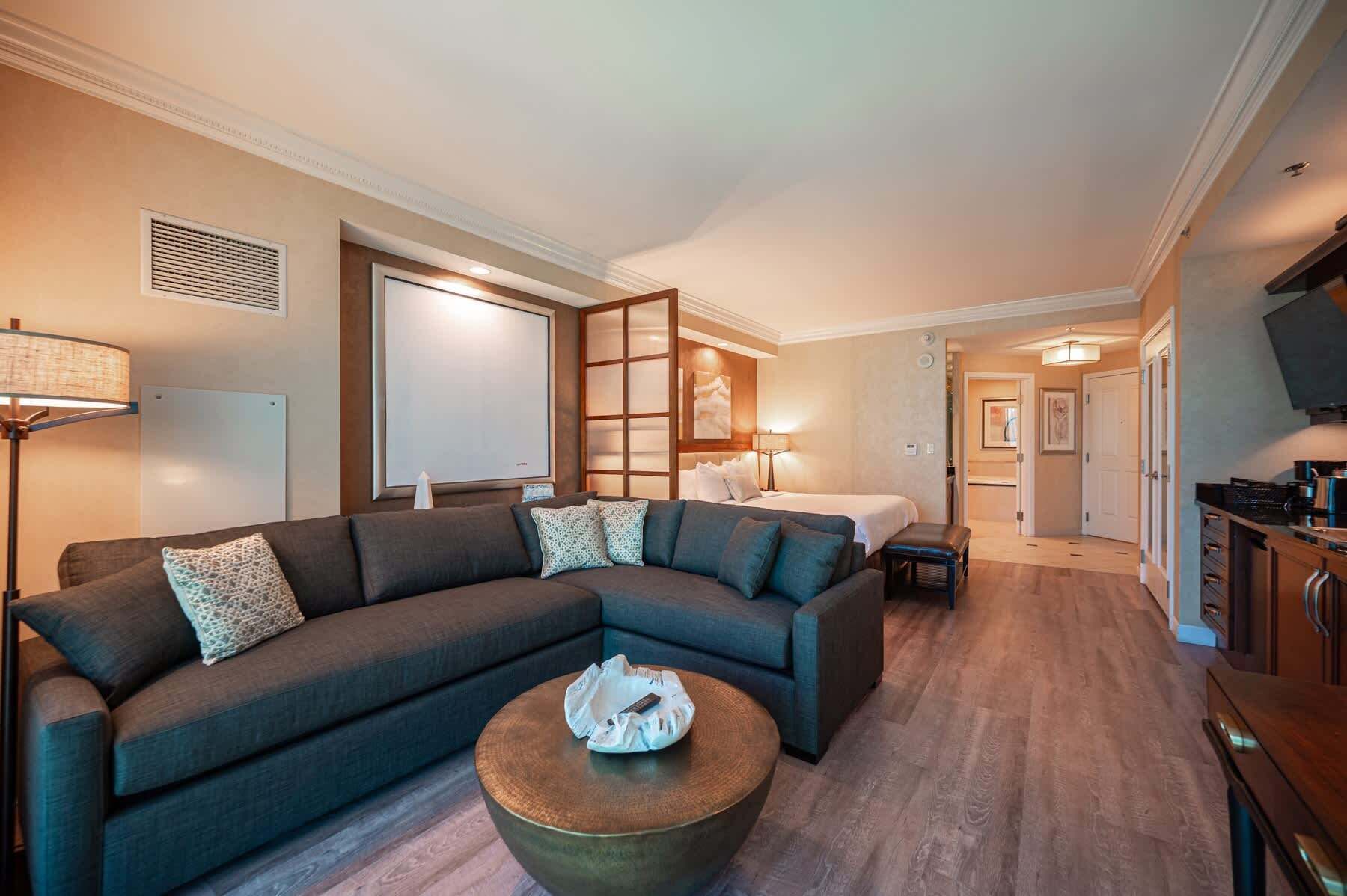 A modern living room with a sectional sofa, a round coffee table, and a bed in the background.
