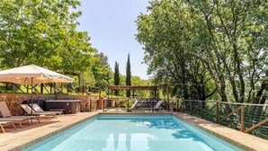 Ein rechteckiger Pool mit Fliesenrand, umgeben von einer Holzterrasse. Liegestühle und ein Grill befinden sich unter einem Sonnensegel.