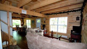 Ein gemütliches Blockhaus-Interieur mit einer großen beigefarbenen Couch, einem Holzespeisesaal und Stühlen. Ein Fernseher steht auf einem Holzschrank, und der Raum hat einen rustikalen Charme mit Holzbalken und einem großen Fenster.