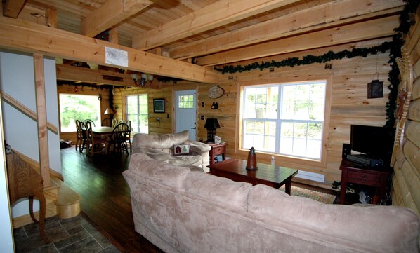 L'intérieur d'un chalet confortable, avec un grand canapé beige, une table à manger en bois et des chaises. Un téléviseur est posé sur un meuble en bois, et la pièce dégage un charme rustique grâce à ses poutres apparentes et à sa grande fenêtre.