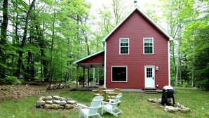 Ein rotes Holzhaus mit Veranda, umgeben von einem Wald. Auf der Wiese stehen Adirondack-Stühle und ein Grill.