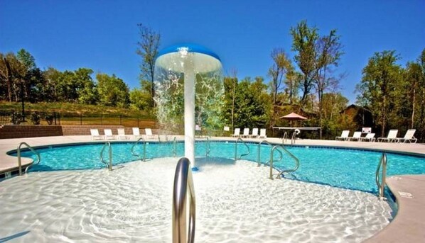 Una piscina con un elemento acuático en el centro, rodeada de sillas de descanso y árboles.