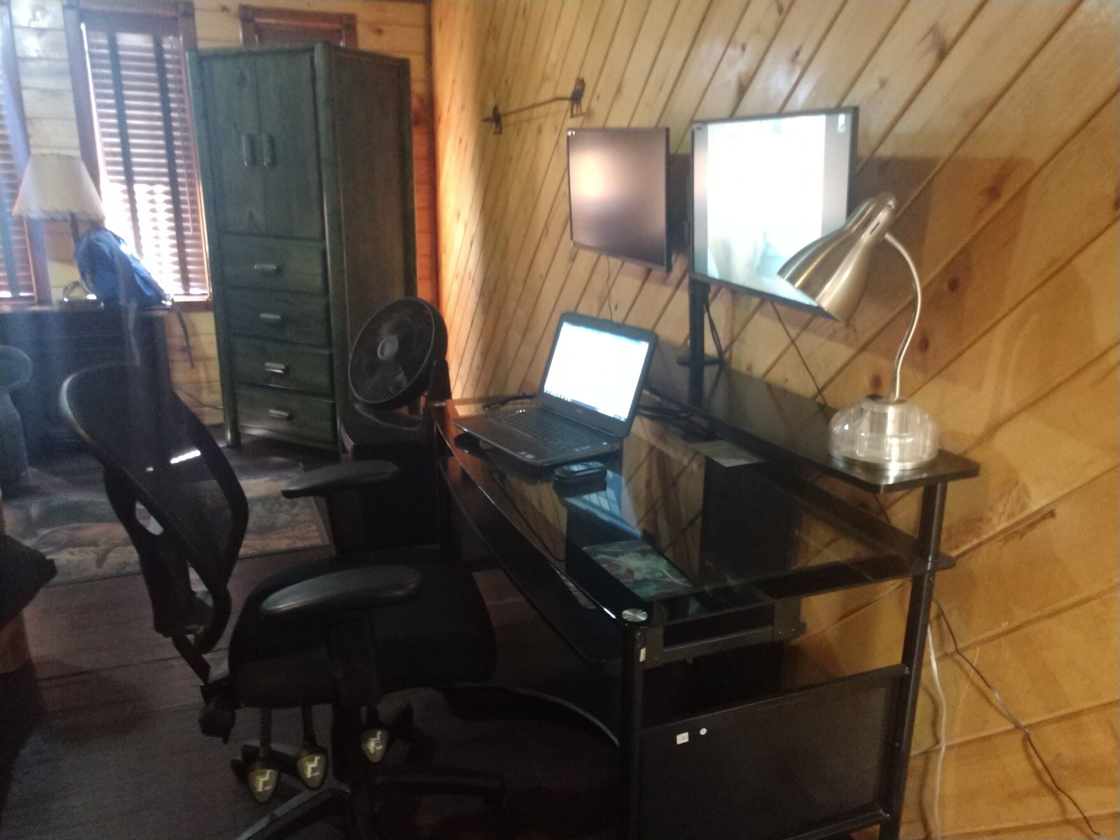 Een kamer met houten inrichting, een bureau met een laptop, een stoel, een ventilator en een kast.