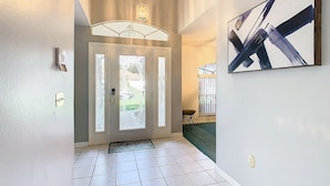 A hallway with a white tiled floor, a glass door with a frosted pattern, a hanging light fixture, and a framed abstract painting on the wall. - ALWAYS FREE POOL HEAT - 2 KINGS, Games room! 5 smart TV’s - S/Steel appliances (Kissimmee)