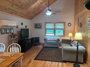 A cozy living room with a wooden ceiling, a television, a fireplace, and a window with a view of trees.