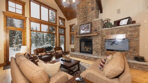 A spacious living room with large windows, a stone fireplace, and a television mounted on the wall. - Last Nickel Lodge: Private Home on Golf Course, Hot Tub, Pool Table, Foosball (Breckenridge)
