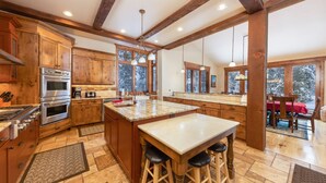 A spacious kitchen with wooden cabinets, a large island with stools, and a view of snowy trees through the windows. - Last Nickel Lodge: Private Home on Golf Course, Hot Tub, Pool Table, Foosball (Breckenridge)