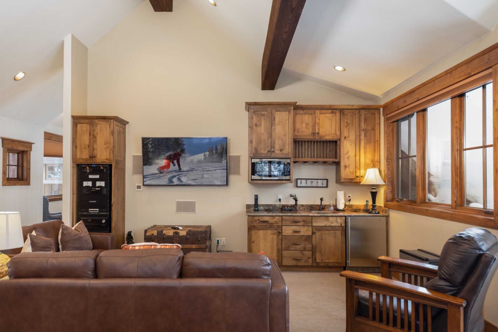 A spacious living area with a brown leather sofa, wooden cabinets, a mounted TV, and a window with a view.